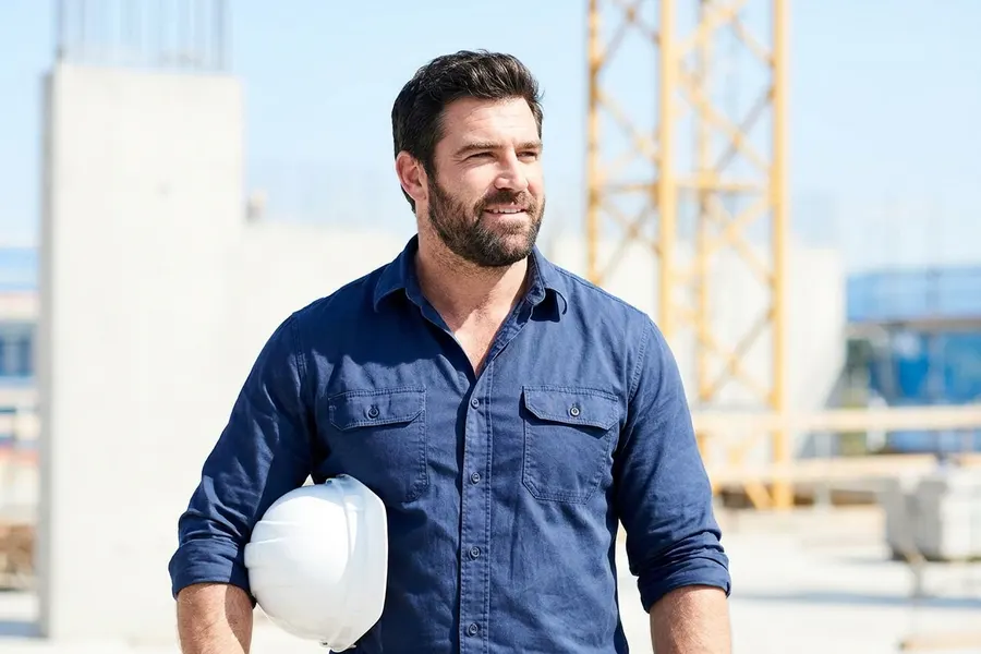 A man in a work shirt on a sunlit building site, hard hat under one arm