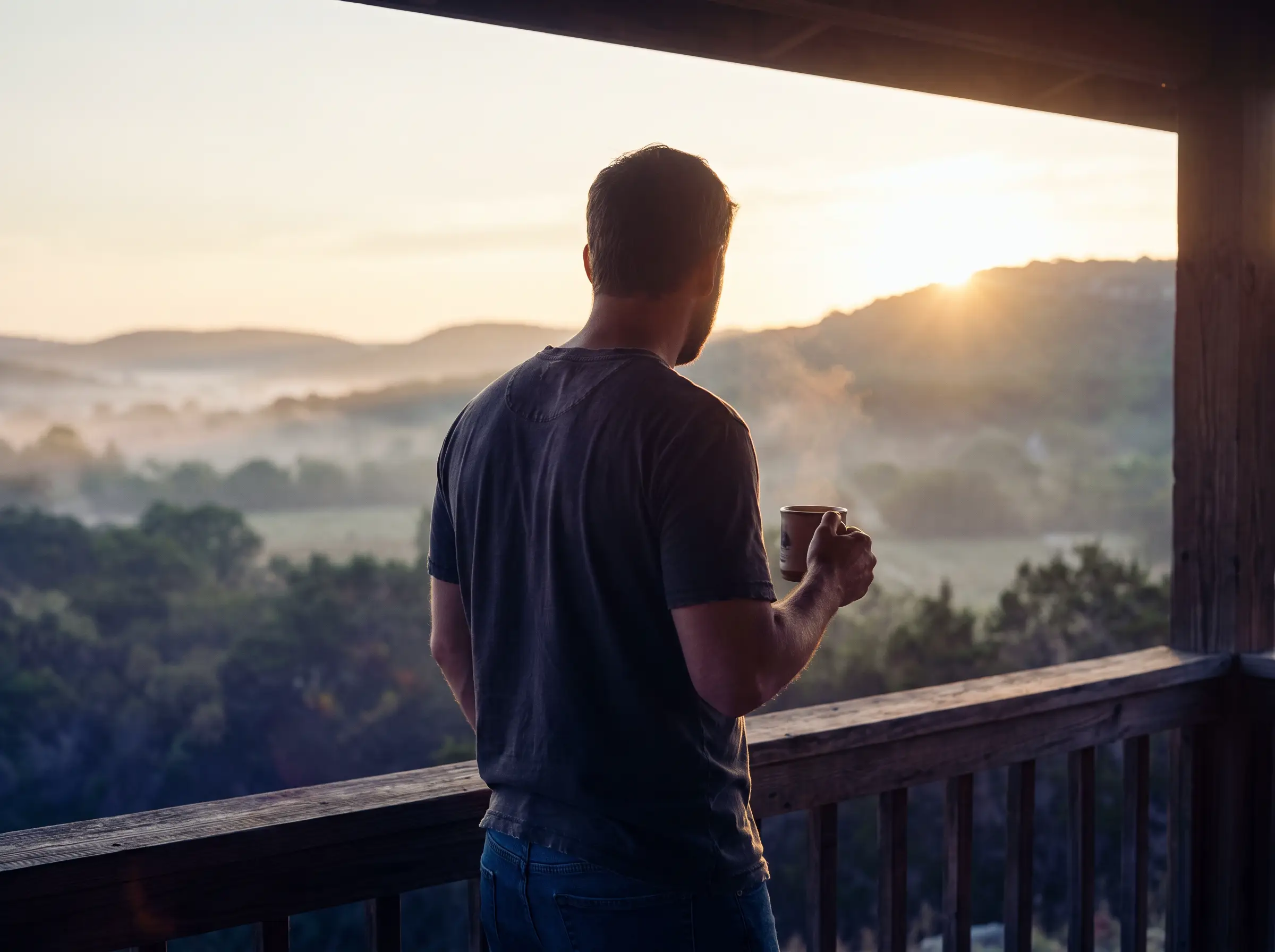 A man on a ranch porch at dawn in the Texas hill country, looking toward the sunrise
